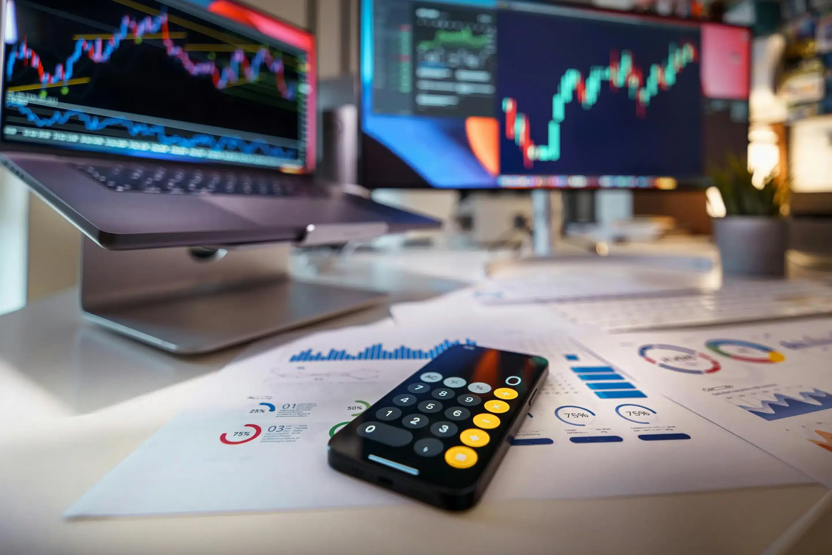 Dermatology Revenue Cycle Management: A Guide A desk with a laptop and computer monitor displaying graphs, with papers and a phone on the desk displaying different numbers, charts, and graphs.