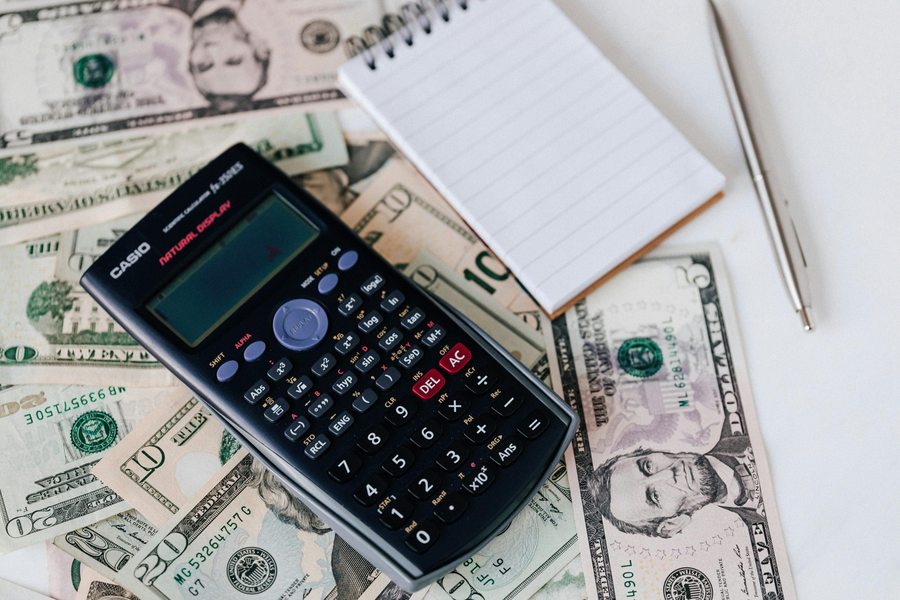 A table covered in money bills with a notepad, pen, and calculator on top.